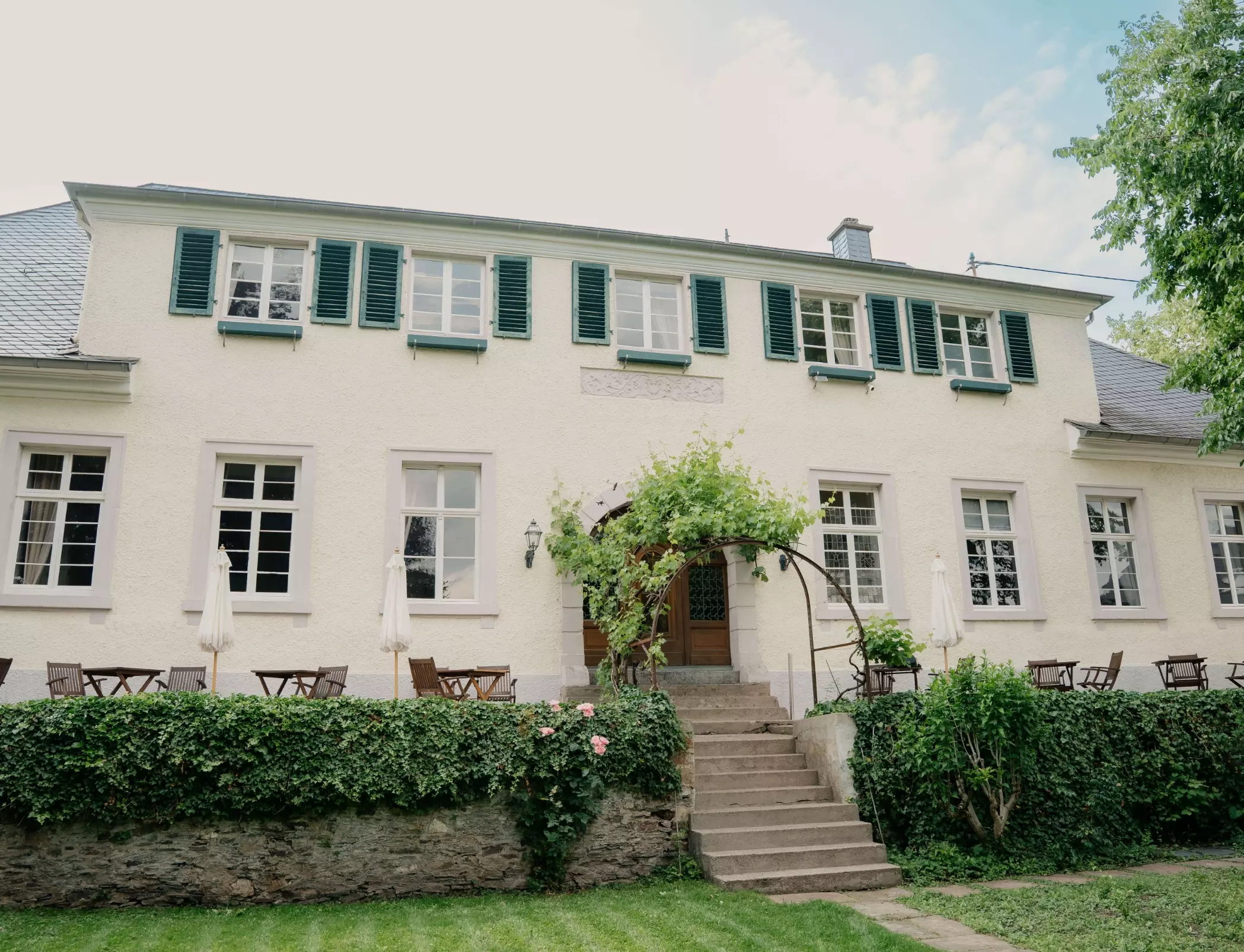 Rückansicht des Hotel Karolingerhofs vom Garten mit weißer Fassade, grünen Fensterläden, Sonnenterrasse, Steintreppe und gepflegten Grünanlagen